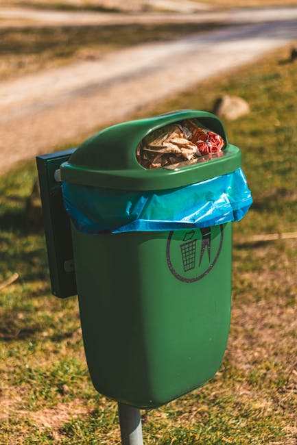 A green outdoor waste bin with a semi-transparent lid containing plastic and paper waste, positioned on a grassy area with a pathway in the background. The platform of the bin features a black symbol 